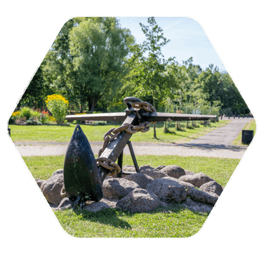 A large outdoor sculpture. The sculpture is of a large anchor with a rusted chain wrapped around it and the anchor is sat on a bed of large rocks 