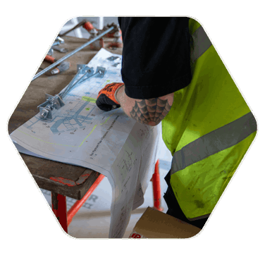 A work mans hands and arms pictured leant over a table showing blueprints to a building. The man is clearly wearing a high vis jacket and has heavy duty gloves on his hands