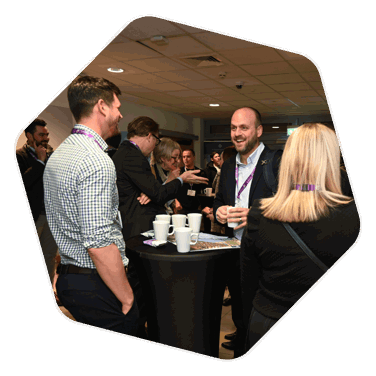 A networking event, a room full of people and in the forefront of the image are two men conversing over a poseur table