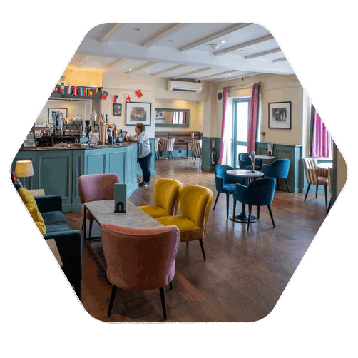 A brightly coloured bar in a hotel, there are tables and chairs as well as a bar with stools situated along the edge of the wooden bar. A lone lady stands at the bar leaning onto the wood.