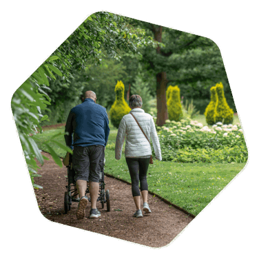 Man and a woman walking on a dirt track with grass and trees around them, pushing another person in a wheelchair