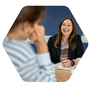 A woman smiling in a meeting room , speaking to another person. The woman has a pen in hand poised to write in a notepad at her hand level on the table
